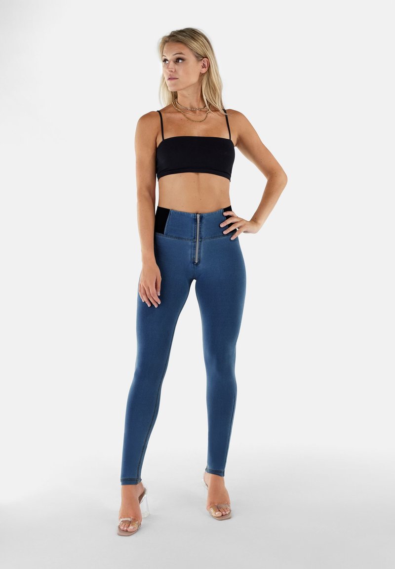 Leggings in denim blu alti in vita con zip frontale, cintura nera e pannelli laterali, abbinati a un top nero corto.