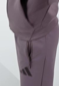 Pantalones deportivos de color morado claro con cintura ajustable mediante cordón, bolsillos laterales y acentos de logotipo negro en la pierna izquierda. Textura de tejido suave.