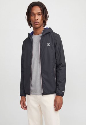 Jeune homme avec des dreadlocks portant une veste à capuche noire non fermée sur une chemise grise et un pantalon blanc cassé, debout devant un fond uni.