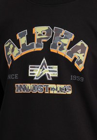 Felpa nera con il bold "ALPHA" in evidenza, dettagli in camouflage, bordature arancioni e la parola "INDUSTRIES" in bianco. Anno "1959" stampato in grigio.