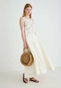 Witte katoenen maxi-jurk met ingewikkelde zilveren borduursels op de buste. Model houdt een ronde, geweven strooien tas vast en draagt zwarte sandalen.