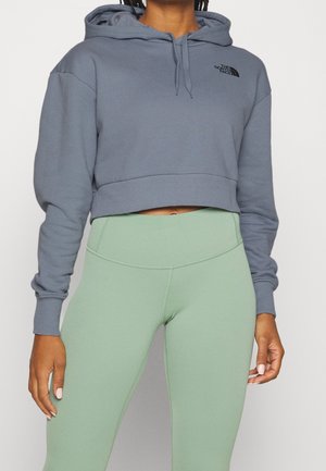Osoba nosząca krótki szary kaptur z logo The North Face i jasnociemnozielone legginsy z wysokim stanem na jednolitym tle.