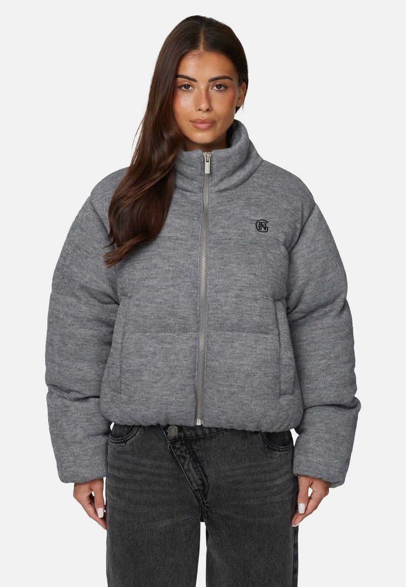 Wygodna szara kurtka z wysokim kołnierzem, zamkiem błyskawicznym z przodu i logo. Posiada szerokie, pikowane sekcje oraz długie rękawy. Noszona z ciemnymi dżinsami.