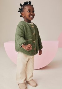 Grön quiltad jacka med dekorativ sömnad, beige ribbade byxor och mjuka bruna tofflor. Rund rosa bakgrund framhäver outfiten.