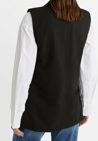 Schwarze ärmellose Weste mit strukturiertem Design, kombiniert mit einem weißen Langarmhemd und blauen Jeans. Glatter Stoff mit minimalen Akzenten.