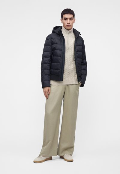 Jeune homme portant une doudoune noire par-dessus un pull torsadé beige avec un pantalon large beige et des chaussures claires.