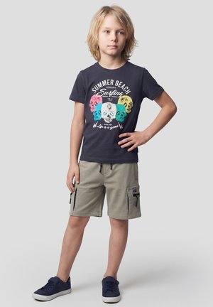 Niño rubio de pie con la mano en la cadera, vistiendo una camiseta azul marino con calaveras coloridas y pantalones cortos beige con bolsillos con cremallera.