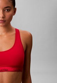 Rotes Sport-BH mit glatter Textur, dünnen Trägern und einem schwarzen Calvin Klein-Logo entlang des Bundes, vor einem neutralen Hintergrund getragen.
