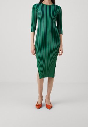 Kobieta ubrana w zieloną sukienkę midi z czarnymi pionowymi paskami i rękawami o długości 3/4, zestawioną z pomarańczowymi szpiczastymi szpilkami.