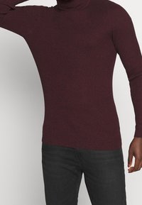 Burgunderfarbener gerippter Rollkragenpullover mit langen Ärmeln, taillierter Form und strukturierter Stoffqualität. Getragen mit schwarzen Hosen, die einen dezenten Glanz aufweisen.