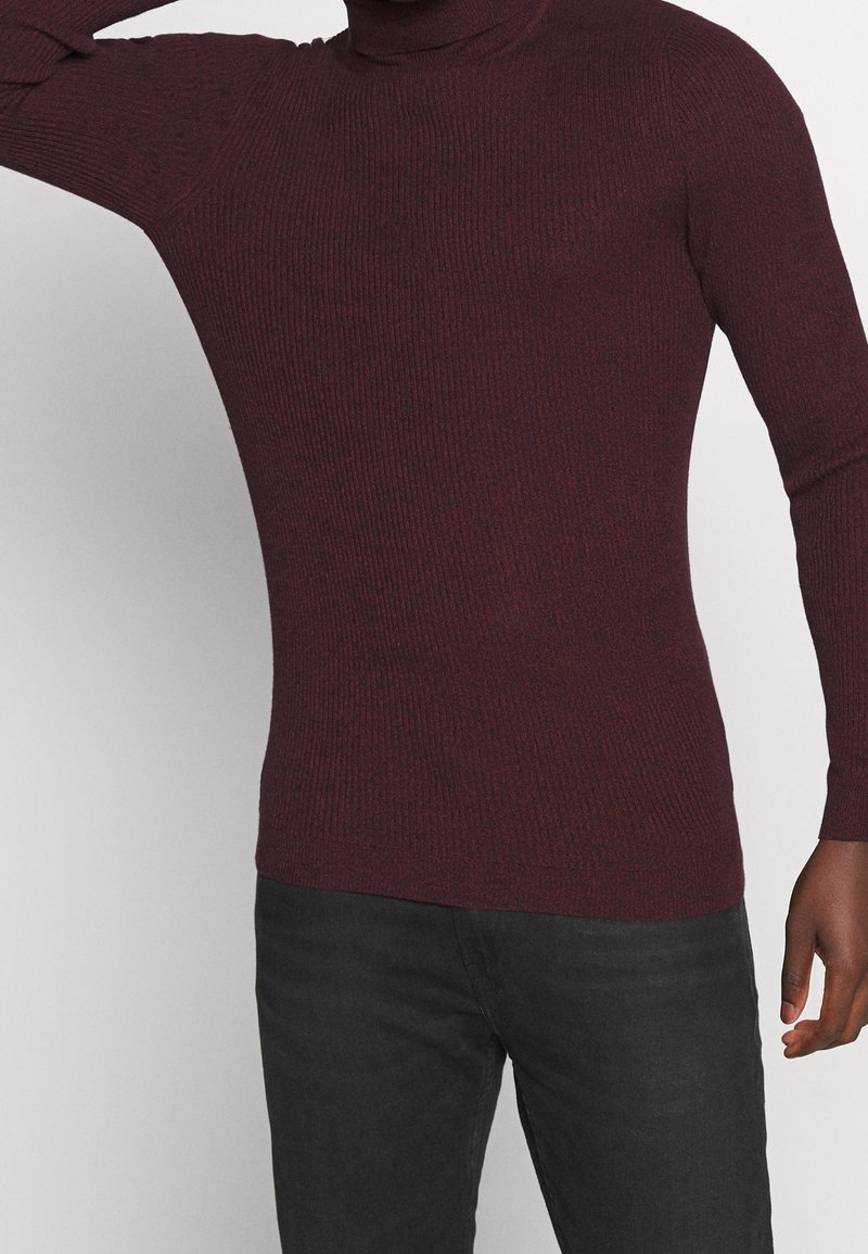 Burgunderfarbener gerippter Rollkragenpullover mit langen Ärmeln, taillierter Form und strukturierter Stoffqualität. Getragen mit schwarzen Hosen, die einen dezenten Glanz aufweisen.
