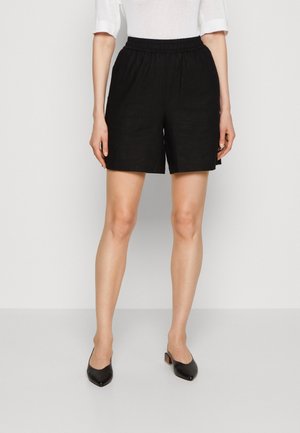 Femme portant un short à taille élastique noir, un haut blanc à manches mi-longues et des chaussures noires à talons bas et bouts pointus, debout sur un fond uni.