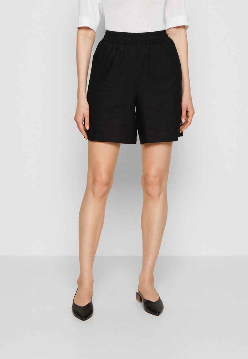 Femme portant un short à taille élastique noir, un haut blanc à manches mi-longues et des chaussures noires à talons bas et bouts pointus, debout sur un fond uni.