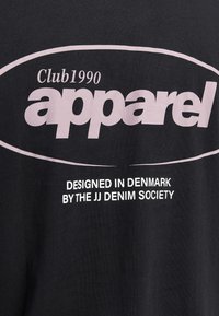 Svart bomull t-skjorte med stor rosa tekst som sier "Club1990 klær" og mindre hvit tekst under, som indikerer designet av JJ Denim Society.