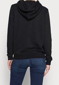 Sudadera negra de material suave, con capucha ajustable mediante cordón, mangas largas y bajo acanalado. Combinada con jeans azul oscuro.