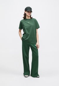 Γυναίκ α με σκούρο πράσινο μπλουζάκι Nike, ταιριαστά φαρδιά παντελόνια, μαύρο καπέλο και γκρίζα αθλητικά, που στέκεται σε λευκό φόντο.