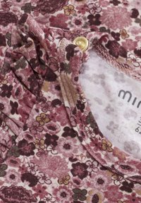 Blommönstrad tyg i nyanser av rosa och burgundy, med en lätt texturerad yta, samt en guldfärgad knapp nära halsringningen.