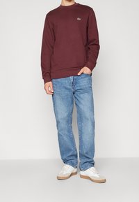 Hombre vestido con sudadera de manga larga burdeos con un pequeño logo verde, jeans azul claro, zapatillas blancas con suelas marrón claro, mano en el bolsillo.
