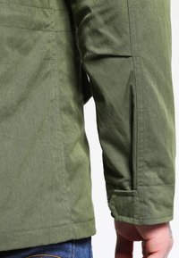 Grön, texturerad jacka i tyg med en figurnära silhuett, sömdetaljer och en manschett med knappstängning.