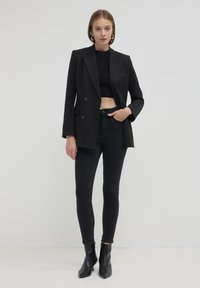 Blazer nero doppiopetto con un design strutturato, abbinato a un crop top nero e jeans neri aderenti, completato da stivali alla caviglia con tacco appuntito.