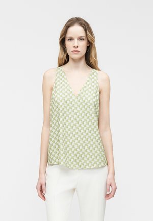 Žena stojaca pred jednoliatym pozadím, oblečená v bezrukávovom zelenobielom top s výstrihom do V a nohaviciach bielej farby s károvaným vzorom.