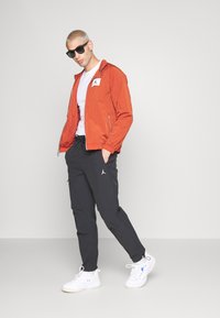 Orangene Windjacke mit Reißverschluss und Logo-Patch, kombiniert mit schwarzen Jogginghosen mit Logo-Details und weißen Sneakers. Sonnenbrille getragen.