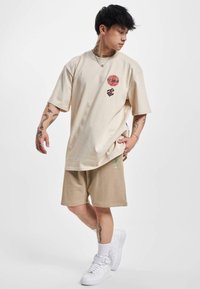 Beige, túlnőtt póló piros kör alakú logóval és barna felirattal, khaki sorttal és fehér sportcipővel párosítva. Dekoratív pálma részlet a sorton.