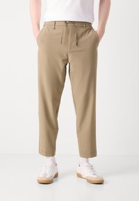 Beige chinos ar auklu jostas, sašaurinātiem svārkiem un sānu kabatām. Valkāti ar baltām snekeriem un potīšu garuma zeķēm. Gluda auduma tekstūra.