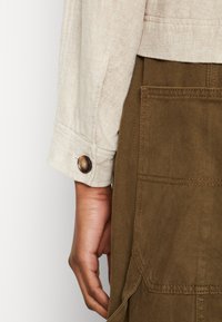 Beige textuur jacket mouw met een knoopdetail, gecombineerd met een donker olijfgroen stof met zakken en zichtbare stiksels.