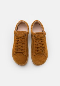 Barna velúr sportcipő, lekerekített orral és lapos fűzőkkel. Texturált felsőrésze és kontrasztos gumikoptatott talpa van. Tiszta dizájn.