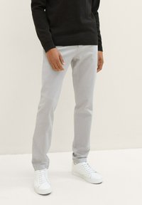 Man in een lichtgrijs broek, zwart sweatshirt en witte veterschoenen staat tegen een effen lichte achtergrond.