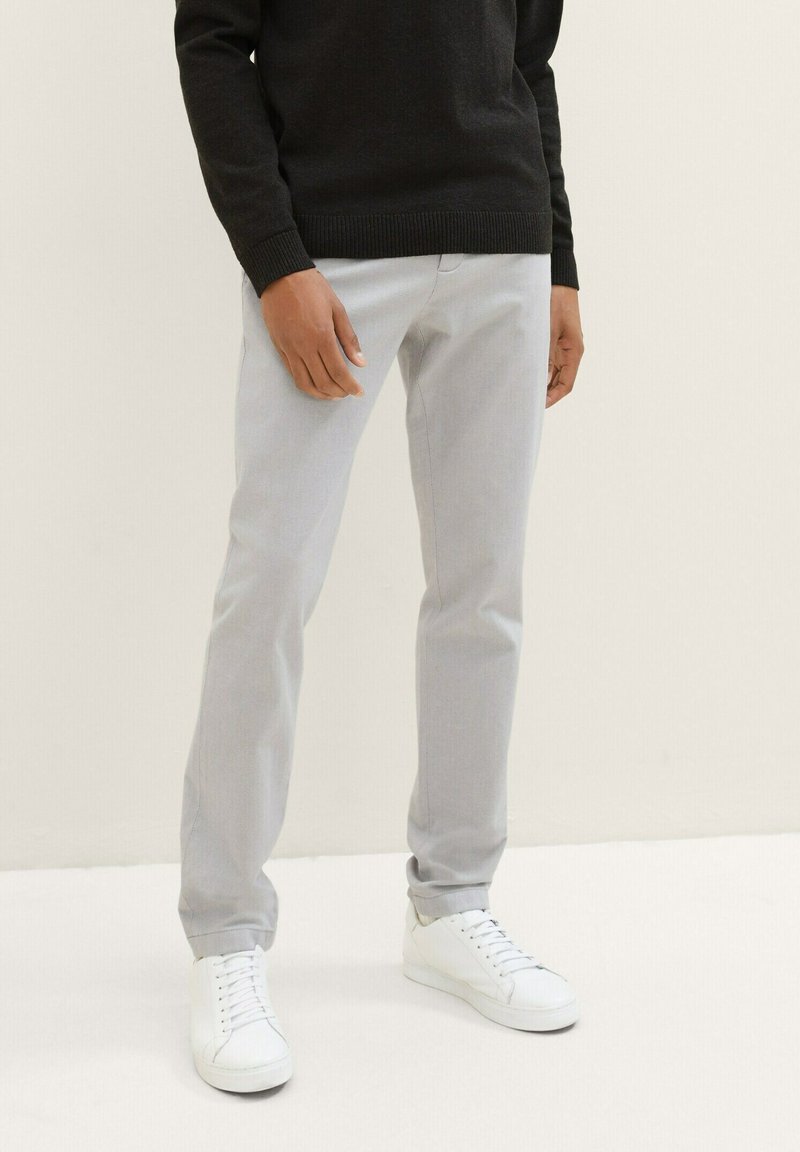 Man in een lichtgrijs broek, zwart sweatshirt en witte veterschoenen staat tegen een effen lichte achtergrond.
