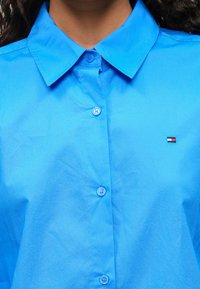 Hele sinine nööpidega särk klassikalise kraega, millel on väike broderingutega logo, kaks nööpi ja sile tekstuur.