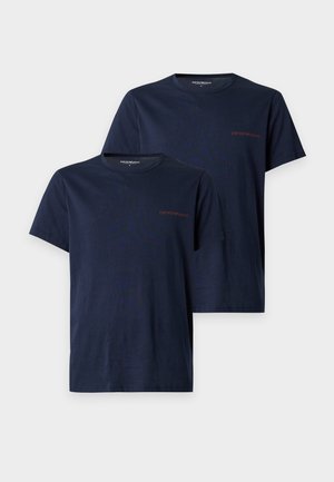 Ein Mann steht barfuß, trägt ein dunkelblaues T-Shirt und weiße Unterwäsche vor einem einfarbigen weißen Hintergrund.