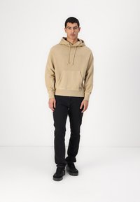 Felpa beige con tasca a marsupio, cordini e logo in rilievo. Abbinata a jeans neri e sneakers nere. Texture liscia.