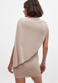 Robe drapée beige en tissu léger et doux. Présente un design asymétrique, sans manches, et un dos fluide.