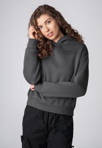 Donkergrijze hoodie met een gestructureerde afwerking, voorzien van een bedrukt label op de borst en geribde boorden en zoom. Gecoördineerd met zwarte broeken.