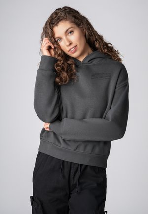 Dunkelgrauer Kapuzenpullover mit strukturierter Oberfläche, ausgestattet mit einem bedruckten Etikett auf der Brust sowie gerippten Bündchen und Saum. Kombiniert mit schwarzen Hosen.