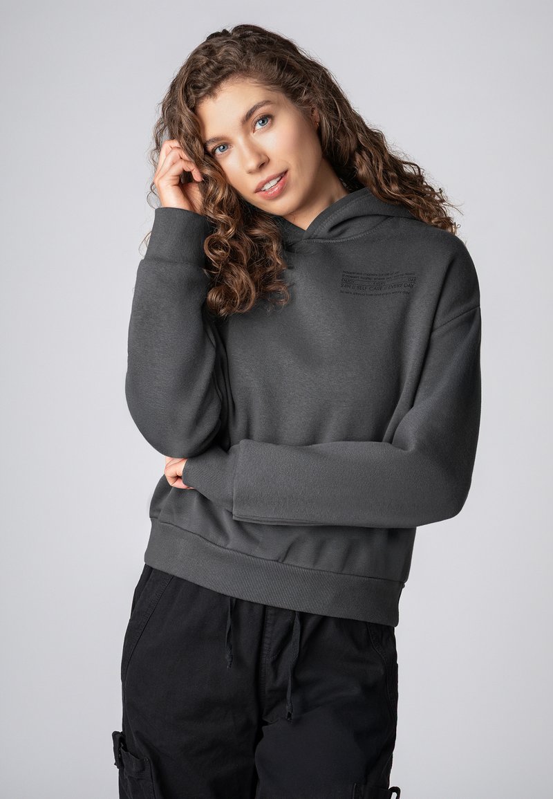 Donkergrijze hoodie met een gestructureerde afwerking, voorzien van een bedrukt label op de borst en geribde boorden en zoom. Gecoördineerd met zwarte broeken.