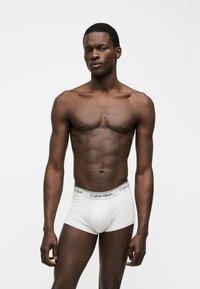 Άνδρας μοντέλο με αθλητική σωματική διάπλαση, φορώντας λευκά μποξεράκια Calvin Klein, μπροστά σε απλό ανοιχτό γκρι φόντο.