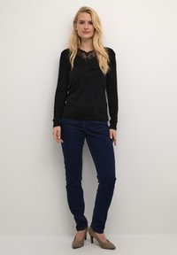 Top nero a maniche lunghe con dettagli in pizzo sul collo, abbinato a jeans blu scuro e tacchi beige. Texture liscia e design aderente.