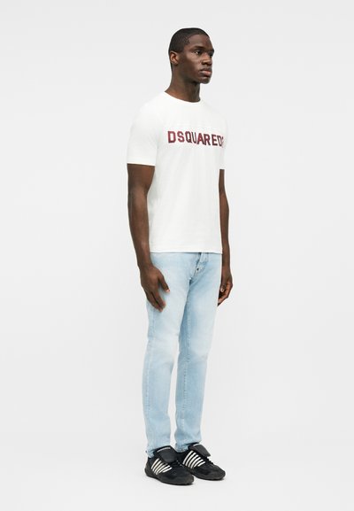 Άνδρας που φοράει λευκό μπλουζάκι DSQUARED2, ανοιχτόχρωμα μπλε τζιν και μαύρα αθλητικά παπούτσια, όρθιος μπροστά σε απλό λευκό φόντο.