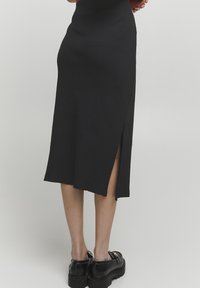 Schwarzer gerippter Midirock mit seitlichem Schlitz, ausgestattet mit einem taillierten Bund und einem geraden Schnitt. Tragen mit klobigen schwarzen Schuhen.