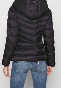 Sort pufferjakke med hætte, der har et quiltet design, horisontale sømme og en glat, blank overflade. Kombineret med blå jeans.