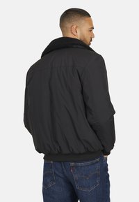 Giubbotto bomber nero con collo in pelliccia sintetica, polsini e orlo elasticizzati, caratterizzato da una texture liscia e una vestibilità comoda. Indossato con jeans blu.