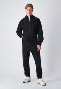 Sudadera negra con cremallera y cuello alto, combinada con joggers a juego. Ambos están hechos de una tela suave que presenta sutiles bolsillos con cremallera.