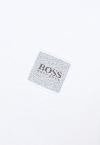 Valkoinen kangas, jossa on röpelöinen neliölaastari, jossa on harmaa kuvio, painettu "BOSS" ja "HUGO BOSS" -teksti tummanruskeana.