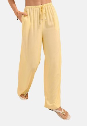 Kvinde iført løse, lysegule bukser med snøre i taljen og sidelommer, kombineret med hvide thongsandaler med gyldne detaljer.