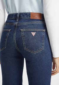 Vaqueros de mezclilla azul oscuro de cintura alta, con una etiqueta de cuero marrón y un logo rojo en forma de triángulo en el bolsillo trasero. Costuras en contraste visibles.