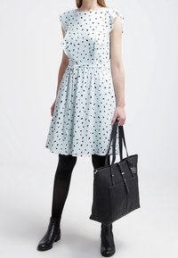 Femme portant une robe blanche à pois noirs, des collants noirs, des bottines noires et tenant un grand sac fourre-tout en cuir noir.
